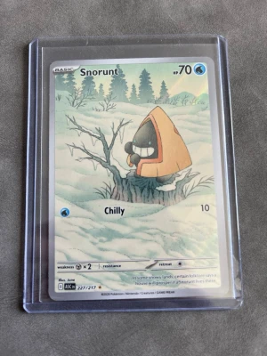 Snorunt Pokémonkort 227/217 - Säljer ett Snorunt Pokémonkort med nummer 227/217 från Scarlet & Violet-serien. Kortet har en vinterinspirerad illustration med Snorunt i orange huva, HP 70 och attacken Chilly. Kortet är i plastskydd och har klassisk Pokémon-design på baksidan.