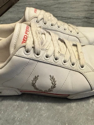 Vita Fred Perry sneakers med röd detalj - Snygga vita sneakers från Fred Perry med klassisk lagerkrans-logga på sidan och röda detaljer på plösen och sulan. Skorna har lågt skaft, vita snören och är tillverkade i slätt läder. Perfekta för en clean och stilren look.
