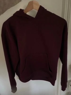 Mörkröd hoodie med huva - En enkel och stilren mörkröd hoodie med huva och stor magficka framtill. Hoodien har långa ärmar och ribbade muddar vid ärmslut och nederkant. Perfekt för att chilla eller hänga med kompisar. Materialet känns mjukt och bekvämt.