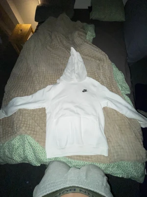 Vit Nike hoodie med logga - Vit hoodie från Nike med klassisk svart logga på bröstet. Tröjan har en stor huva, ribbade muddar och en praktisk magficka. Perfekt för dig som gillar sportig och clean stil. Materialet är mjukt och bekvämt, passar till chill och streetwear.