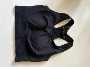 Svart sport-bh från Röhnisch - Snygg svart sport-bh från Röhnisch med brottarrygg och bred resår nedtill. Tillverkad i stretchigt funktionsmaterial som ger bra stöd vid träning. Diskret logga på resåren och sömlös design för extra komfort.