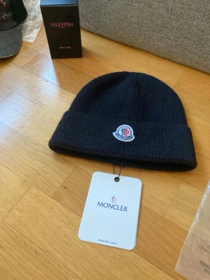 Svart Moncler mössa med logga - Säljer en svart ribbstickad mössa från Moncler med uppvikt kant och Monclers klassiska logotyp framtill. Mössan är tillverkad i mjukt material och har en stilren, enkel design som passar perfekt till höst och vinter.Man får med tagg och påse till 