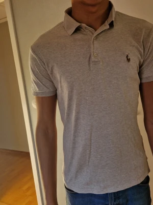 Ljusgrå polo t-shirt från Ralph Lauren - Säljer en ljusgrå polo t-shirt från Ralph Lauren med klassisk krage och knappslå framtill. T-shirten har korta ärmar och den ikoniska broderade loggan på bröstet. Perfekt för dig som gillar stilrena och tidlösa plagg. Säljer pga att den är flr liten för mig 177 cm lång.