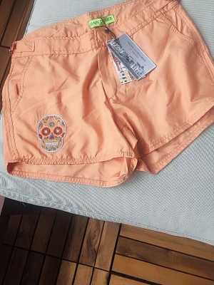 Persikofärgade strandshorts - Snygga persikofärgade strandshorts från Bain de Mer med broderad dödskalle på ena benet. Shortsen har fickor, justerbara spännen i midjan och är tillverkade i ett lätt och snabbtorkande material – perfekta för stranden! Midjemått:39 cm.