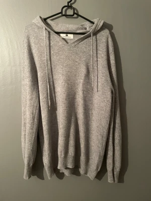 Ljusgrå hoodie från Drakenberg - Mysig ljusgrå hoodie från Drakenberg med huva och dragsnören. Tillverkad i mjukt stickat material som känns skönt mot huden. Klassisk passform med ribbade muddar vid ärmslut och nederkant. Perfekt för en avslappnad och stilren look.