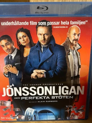 Jönssonligan Den Perfekta Stöten Blu-ray - Säljer en svensk actionkomedi på Blu-ray, Jönssonligan Den Perfekta Stöten. Fodralet är blått och filmen är regisserad av Alain Darborg. Omslaget har en röd bakgrund med bilder på huvudkaraktärerna och detaljer som pengar och ett kassaskåp. Speltid: 92 min, rekommenderas från 11 år.