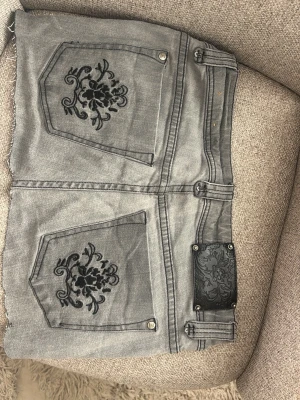 Grå jeanskjol med broderi - Säljer en grå jeanskjol, snygga svarta broderade detaljer på bakfickorna. Perfekt för dig som gillar en edgy och unik stil.