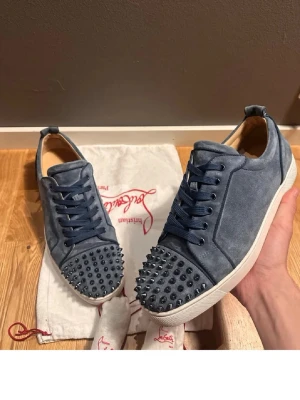 Blåa sneakers med nitar från Louboutin - Snygga blåa sneakers från Christian Louboutin i mocka med coola nitar på tån. Skorna har klassisk låg profil, blåa skosnören och vit sula. Perfekt för dig som vill sticka ut med en edgy och lyxig stil.