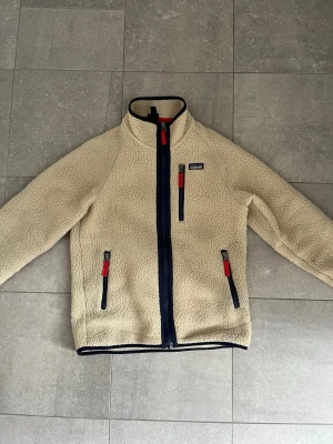 Beige teddyjacka från Patagonia - Säljer en beige teddyjacka från Patagonia med marinblå dragkedjor och röda detaljer. Jackan har hög krage, tre fickor med dragkedja och är gjord i mjukt, fluffigt teddy-material. Perfekt för dig som vill ha en mysig fleece. Vid frågor elle nåt annat är de bara att höra av sig!
