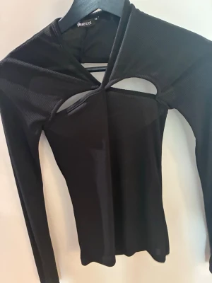 Svart långärmad topp från Gina Tricot - Trendig svart långärmad topp från Gina Tricot med coola cut out-detaljer framtill och baktill. Toppen har en tight passform och är tillverkad i ett ribbat material som ger en snygg struktur. Perfekt för dig som vill sticka ut med en unik design.
