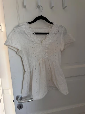 Vit spetsblus med v-ringning - Söt vit blus med v-ringning och spetsdetaljer. Blusen har korta ärmar och är tillverkad i bomull med broderade hålmönster. Perfekt för dig som gillar romantisk och luftig stil.