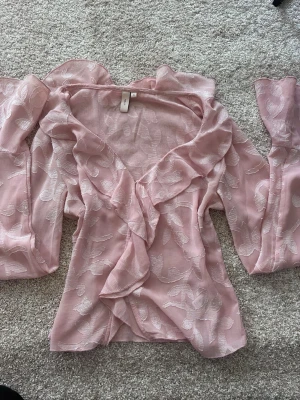 Rosa blus - Söt rosa blus från Nelly med volangkant längs ringningen och framtill. Blusen är transparent med ett blommigt mönster i tyget och har långa, utsvängda ärmar. Perfekt för dig som gillar romantisk och trendig stil. Orginalpris ca 499
