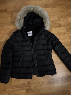Svart dunjacka från Tommy Hilfiger - Snygg svart dunjacka från Tommy Hilfiger med avtagbar huva och fejkpäls. Jackan har quiltad design, dragkedja framtill och två sidofickor. Perfekt för kalla vinterdagar och har en klassisk look med diskret logga på bröstet. Prisset kan diskuteras 