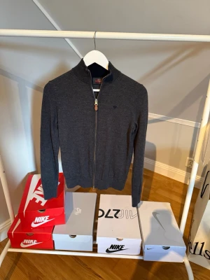 Mörkgrå zip-tröja från Morris - Snygg mörkgrå zip-tröja från Morris i 100% merinoull. Tröjan har hög krage, dragkedja framtill och diskret logga på bröstet. Perfekt för lager-på-lager och håller dig varm under kyliga dagar.