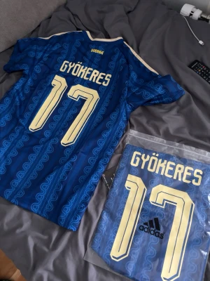Blå Sverige fotbollströja Adidas - Säljer en blå Sverige fotbollströja med Gyökeres och nummer 17 på ryggen. Tröjan har gula detaljer och ett unikt mönster i tyget. Perfekt för dig som vill stötta landslaget med stil.