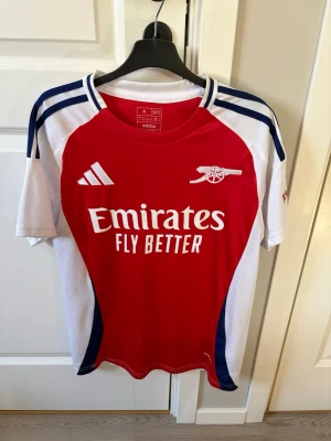 Arsenal matchtröja Adidas röd/vit - Snygg Arsenal matchtröja från Adidas i rött, vitt och marinblått. Tröjan har klubbmärke och Adidas-logga på bröstet samt 'Emirates Fly Better' tryckt framtill. Tillverkad i lätt och ventilerande material, perfekt för fotboll eller träning.