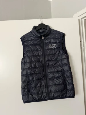Mörkblå dunväst från EA7 Armani - Snygg mörkblå dunväst från EA7 Emporio Armani med quiltad design och hög krage. Västen har dragkedja framtill och logotyp broderad på bröstet. Perfekt för lager-på-lager och ger en sportig look.