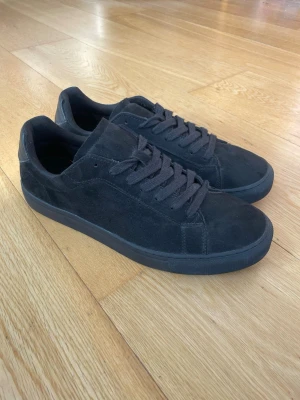Svarta sneakers i mocka - Säljer ett par stilrena svarta sneakers med låg profil och klassisk design. Skorna är tillverkade i mocka med matchande svarta skosnören och gummisula. Perfekta för dig som gillar en clean och enkel look.