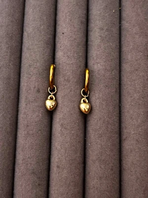 Guldiga örhängen med hjärtberlocker - Snygga små hoops i guldfärg med hängande hjärtformade berlocker. Örhängena har en enkel och stilren design som passar till många olika looks. Materialet ser ut att vara metall med en blank finish. Perfekta för dig som gillar minimalistiska smycken med en söt twist.