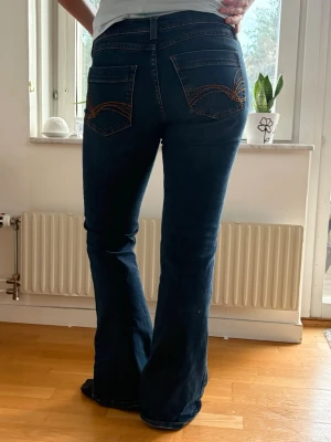 Mörkblå bootcut jeans med brodyr - Mörkblå bootcut jeans med snygg orange brodyr på bakfickorna. Tillverkade i stretchigt denimtyg för skön passform. Perfekta för dig som gillar en retroinspirerad look. Säljer eftersom de inte kommer till användning.