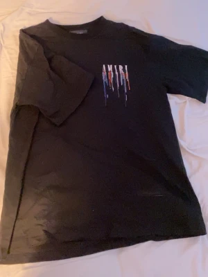 AMIRI T-shirt - Svart t-shirt från AMIRI med tryck av loggan framtill och stort tryck på ryggen. Båda trycken har färgstänk i vitt, blått, orange och rött som rinner ner. Klassisk passform och rund hals. Perfekt för dig som gillar streetwear och vill sticka ut.