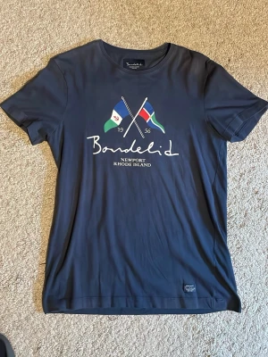 Mörkblå t-shirt Bondelid - Mörkblå t-shirt från Bondelid med tryck av två korsade flaggor och texten 'Newport Rhode Island' framtill. Klassisk rund hals och korta ärmar. Perfekt för en avslappnad stil.