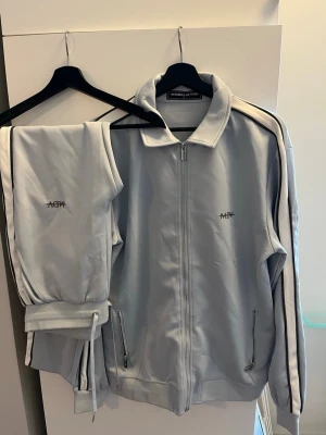 Ljusblå mjukisset från Manière De Voir - Snyggt ljusblå mjukisset från Manière De Voir med gråa detaljer längs sidorna och logga på benet och bröstet. Byxorna har dragsko i midjan och är gjorda i ett mjukt material som känns skönt mot huden. Perfekt för en avslappnad och trendig look. Storlek L