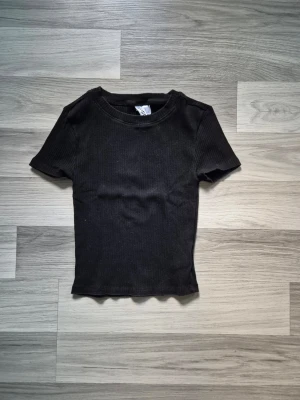 Svart ribbad topp - En svart ribbad topp med korta ärmar och rund hals. Toppen har en slim passform och är tillverkad i mjukt bomullsmaterial. Perfekt för dig som gillar en enkel och stilren look.