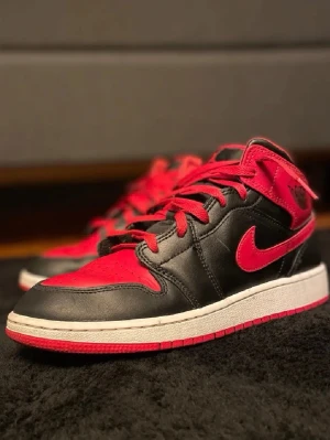 Air Jordan 1 Mid Svart/Röd - Säljer ett par Air Jordan 1 Mid sneakers från Nike i svart och rött läder. Skorna har klassisk Swoosh-logga på sidan, röda snören och en vit/röd sula. Perfekt för dig som gillar ikoniska och sportiga sneakers med cool design.