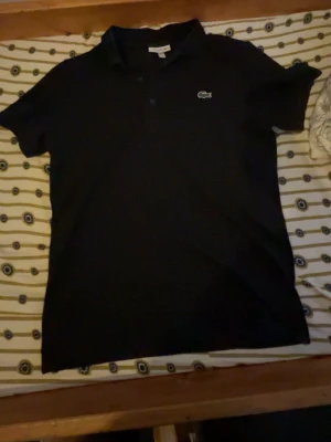 Svart pikétröja från Lacoste - Klassisk svart pikétröja från Lacoste med korta ärmar och den ikoniska krokodilloggan på bröstet. Tröjan har krage och knappar framtill. Perfekt för en stilren och enkel look.storlek m men passar också s