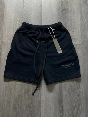 Svarta shorts från Essentials Fear of God - Säljer ett par svarta shorts från Essentials Fear of God med dragsko i midjan och logga tryckt på benet. Sköna och stilrena shorts i mjukt material, perfekta för en avslappnad look.