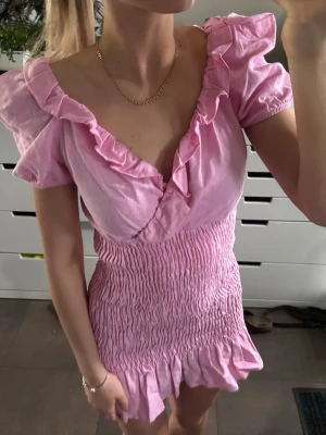 Rosa kortklänning med volang - Supersöt rosa kortklänning med volangdetaljer längs v-ringningen och ärmarna. Klänningen har puffärmar och smockad midja som ger en feminin siluett. Perfekt för sommarens alla tillfällen!