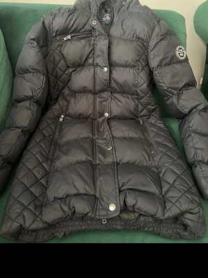 Svart dunjacka från Vinson Polo Club - Snygg svart dunjacka från Vinson Polo Club med quiltade detaljer på sidorna och ärmarna. Jackan har dragkedja och knappar framtill, tre fickor med dragkedja och hög krage. Perfekt för kalla vinterdagar och riktigt varm.