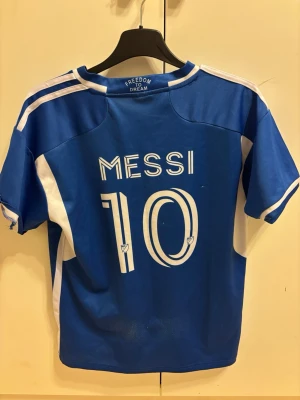 Blå Messi fotbollströja Inter Miami - Säljer en blå fotbollströja från Freedom To Dream med Messi och nummer 10 på ryggen. Tröjan har vita detaljer, korta ärmar och klubbmärke samt sponsortryck på bröstet. Perfekt för dig som älskar fotboll och Messi.
