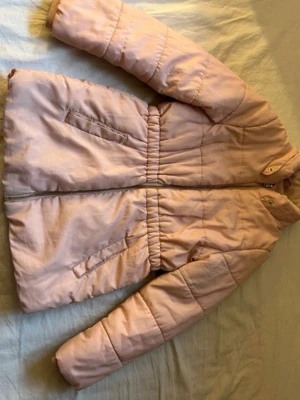 Ljusrosa pufferjacka från H&M för barn  - Ljusrosa pufferjacka från H&M med dragkedja framtill och två snedställda fickor. Jackan har resår i midjan för en snygg passform och är fodrad med mjukt material på insidan. Perfekt för kalla vinterdagar. Finns en lite fläck på jackan se sista bild! 