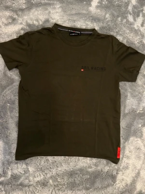 Olivgrön Sail Racing t-shirt storlek M - Olivgrön t-shirt från Sail Racing med svart tryck på bröstet och stor grafisk print på ryggen. Klassisk rund hals och korta ärmar. Tillverkad i mjuk bomull för skön känsla. Storlek M