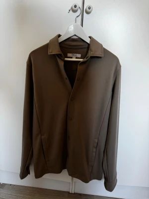 Brun skjorta från SMOG relaxed fit - Stilren brun skjorta från SMOG i relaxed fit. Skjortan har klassisk krage, knäppning framtill och långa ärmar. Perfekt för en chill och avslappnad look. Tillverkad i ett mjukt material som känns skönt mot huden.