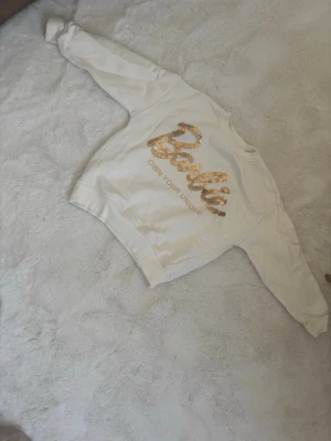 Vit Barbie sweatshirt med paljetter - Cool vit sweatshirt från Barbie med guldiga paljetter som stavar 'Barbie' och texten 'OWN YOUR UNIQUE' på ryggen. Tröjan har rund hals, ribbade muddar och är gjord i mjukt sweatshirtmaterial. Perfekt för dig som gillar statement-plagg och glitter.