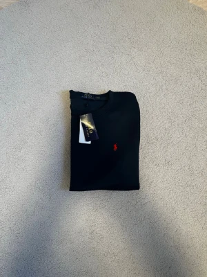 Mörkblå tröja från Polo Ralph Lauren - Snygg mörkblå tröja från Polo Ralph Lauren med klassisk röd logga broderad på bröstet. Tröjan har rund halsringning, långa ärmar och ribbade muddar. Perfekt till jeans eller chinos för en clean look. Väldigt fin är den. Helt ny aldrig använd. Fraktar samma dag eller nästa📦🚚 Aktiv hela tiden✅ hör av dig om du undrar något⁉️