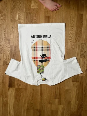 Vit BAPE t-shirt med rutig logga - Vit t-shirt från A Bathing Ape med klassisk BAPE-logga i rutigt mönster och svart tryck. T-shirten har rund hals och korta ärmar. Märkets tag och camo-detalj vid halsen. Perfekt för dig som gillar streetwear.