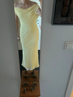 Ljusgul axelbandslös maxiklänning - Snygg ljusgul maxiklänning med off shoulder-design och draperad urringning. Klänningen har öppen rygg med tunna band som knyts i nacken. Perfekt för varma dagar och somriga tillfällen.