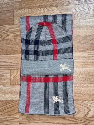 Grå burberry mössa - Snygg grå mössa med klassiskt rutigt mönster i svart, rött och mörkblått. Mössan har en vikt kant och en broderad logga i vitt framtill. Tillverkad i mjukt stickat material som håller dig varm och stilren. Du får även med en halsduk så det blir som ett sett