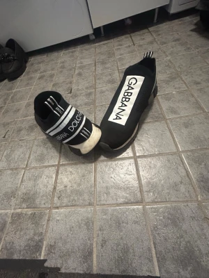 Dolce & Gabbana svarta sneakers - Svarta sneakers från Dolce & Gabbana med tydliga vita logotexter på ovansidan och hälen. Skorna har en strumpliknande passform och elastiskt tyg, samt vit och svart sula. Snygg och modern design som sticker ut.