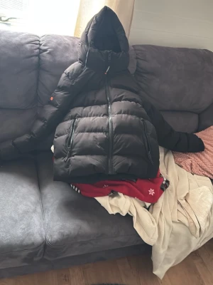 Svart dunjacka från Superdry - Säljer en svart dunjacka från Superdry med huva och två stora dragkedjeförsedda fickor framtill. Jackan har broderad Superdry-logga i orange på ärmen och är riktigt puffig och varm. Perfekt för kalla vinterdagar och har en modern, oversized look.