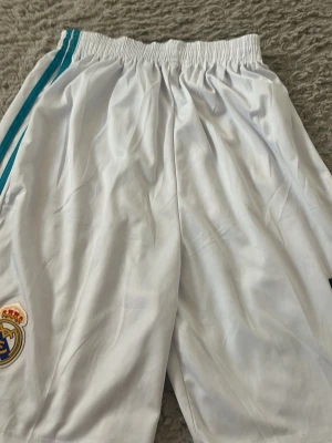 Vita Real Madrid shorts med blå ränder - Säljer ett par vita sportshorts från Real Madrid med klubbens broderade logga på vänster ben och blå ränder längs sidorna. Shortsen har elastisk midja och är tillverkade i ett lätt och svalt material, perfekt för fotboll eller träning.
