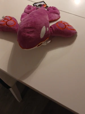 Lila Pokémon Kyogre gosedjur - Säljer ett mjukt och coolt lila Kyogre gosedjur från Pokémon. Gosedjuret har vita och orange detaljer, stora fenor och broderade ögon. Perfekt för Pokémon-fans som vill ha något unikt i samlingen.