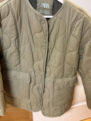 Olivgrön quiltad pufferjacka från Zara - Olivgrön pufferjacka från Zara med vågigt quiltat mönster och rund halsringning. Jackan har tryckknappar framtill och två stora fickor med lock. Materialet är lätt och ger en modern siluett, perfekt för kalla vinterdagar.