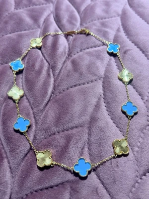 Guldhalsband med blå och guld  klöver - Snyggt halsband med guldfärgad kedja och klöverformade hängen i blått och vitt. Hängena har en elegant pärlad kant och halsbandet har en klassisk karbinhake. Perfekt för dig som gillar färg och unika detaljer.