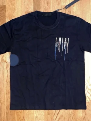 Amiri t-shirt - Snygg svart t-shirt från AMIRI med stort tryck på ryggen och mindre tryck på bröstet. Trycket består av AMIRI-loggan och färgstänk i blått, orange och vitt som rinner ner. Klassisk passform och rund hals. Perfekt för dig som vill sticka ut. Pris går diskuteras vid snabb affär