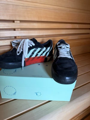Off-White skor - Snygga svarta sneakers från Off-White med vita diagonala ränder på sidan och en röd detalj vid sulan. Skorna har vita skosnören och en unik mönstrad yttersula. Ovandelen är i mocka och textil för en cool streetwear-look.